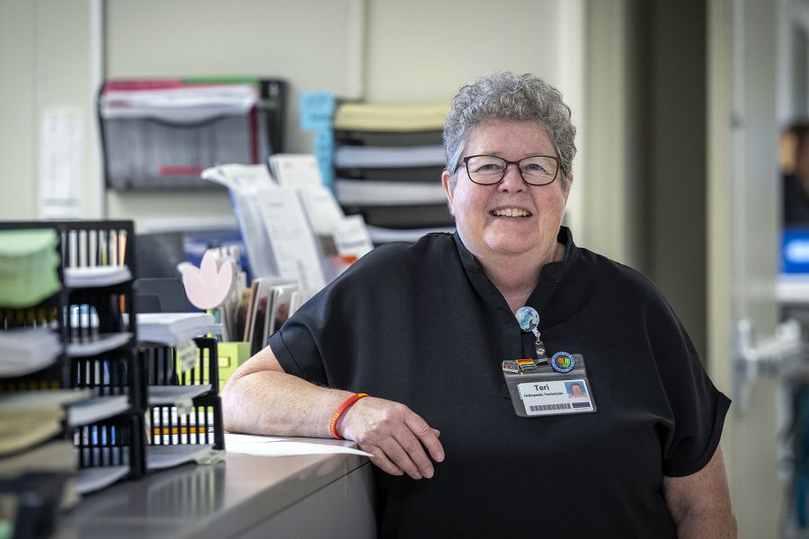 Teri Timpany is leaning against a care desk while smiling directly at the camera. Teri has short, curly, silver hair and wears glasses. She’s wearing black scrubs.