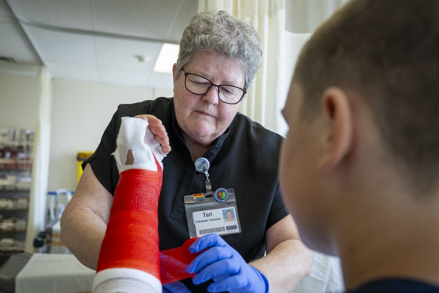 Teri Timpany is putting a red cast on a young patient. She’s looking down at his arm while she puts a cast on him. Teri has short, curly, silver hair and wears glasses. She’s wearing black scrubs.