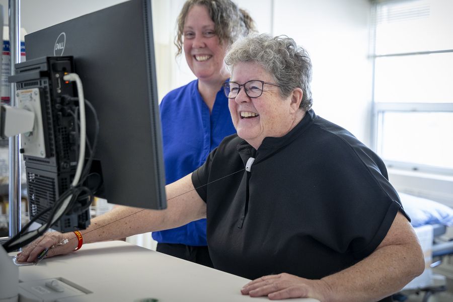 Teri Timpany is standing behind a computer next to a colleague. Both woman are looking and smiling at the screen. Teri has short, curly, silver hair and wears glasses. She’s wearing black scrubs.