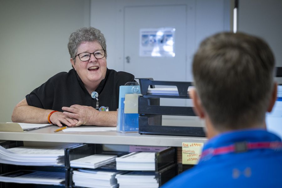 Teri is standing behind a care desk and smiling at a colleague. Teri has short, curly, silver hair and wears glasses. She’s wearing black scrubs.