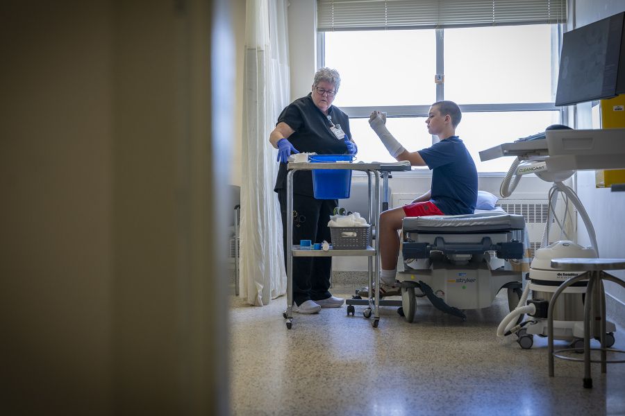 A wide shot of a treatment room where Teri Timpany is standing and a young patient is sitting on a bed. Teri is putting a cast on him.